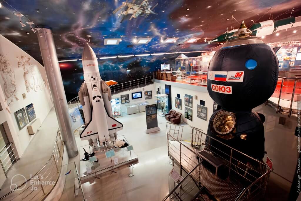 An interior view of the Kosmo Museum in Moscow, showcasing detailed space exhibits, including a model of the Soviet space shuttle "Buran" and a replica of the "Soyuz" spacecraft. The space-themed ceiling enhances the immersive experience.